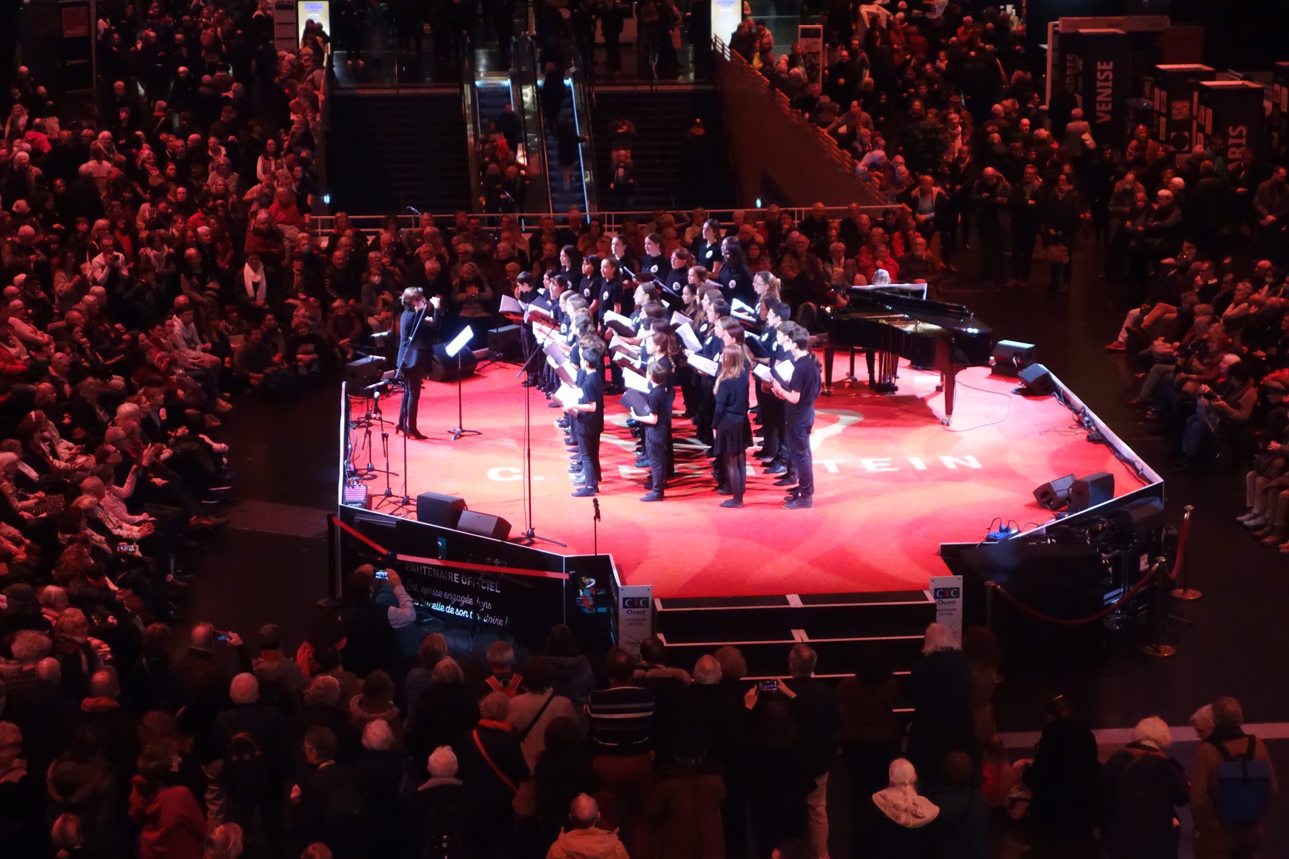 Une cinquantaine d'élèves choristes sur une scène rouge avec leur enseignante à la direction et accompagnés d'une pianiste entourés d'un public nombreux assis dans la pénombre.