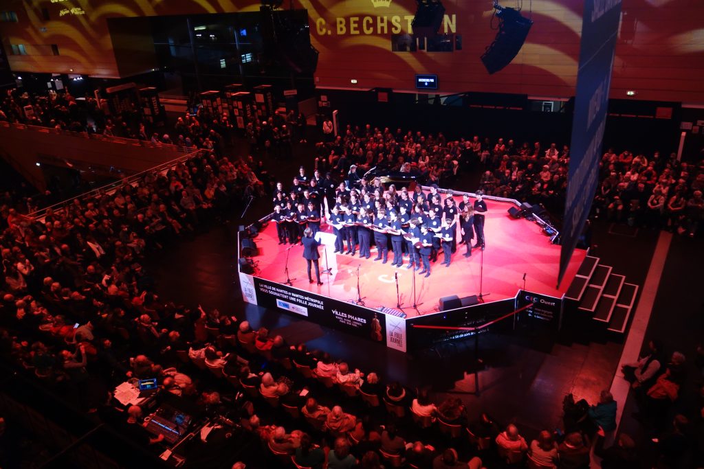 Les élèves des CHAM sur la scène du kiosque de la cité des congrès de Nantes lors de la Folle Journée, entourés par un public assis, sous la direction de Caroline Sorieux et accompagnés par Nathalie Darche au piano.